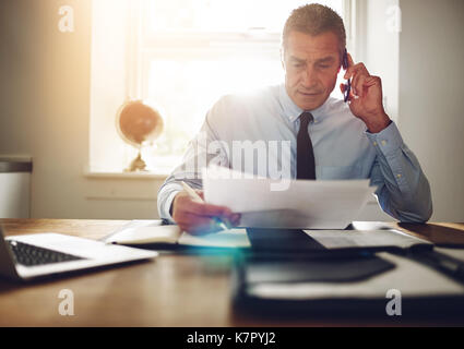 Mature executive discussing documents over the phone while working at his desk in an office Stock Photo