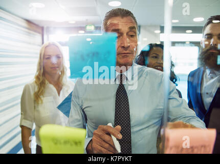 Half body portrait of businessman looking at sticky notes on glass in office with colleagues in background Stock Photo