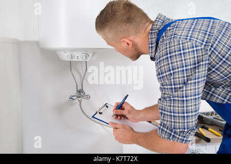 Male Plumber Writing On Clipboard In Front Of Electric Boiler In Kitchen Room Stock Photo