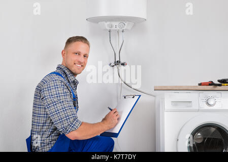 Male Plumber Smiling While Writing Meter Reading Of Electric Boiler On Clipboard Stock Photo