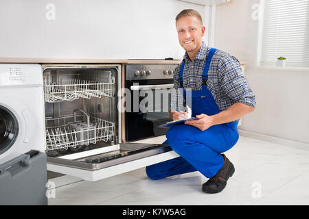 Young Handyman Looking At Dishwasher And Writing On Clipboard Stock Photo