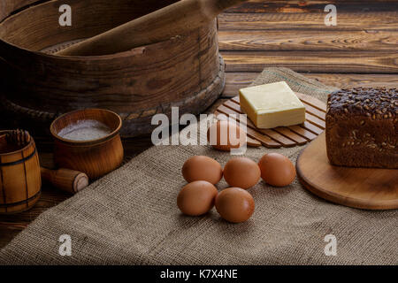 Items and ingredients for baking on a wooden background and burlap. Flour, eggs, rolling pin, whisk, sieve, butter, bread. Stock Photo
