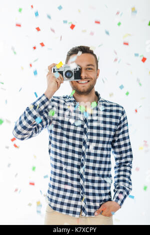 Happy handsome man taking photo with retro film camera and confetti falling on white background. Party concept Stock Photo