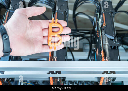 Man hand holds a plasticine bitcoin against the backdrop of a mining farm from video cards Stock Photo