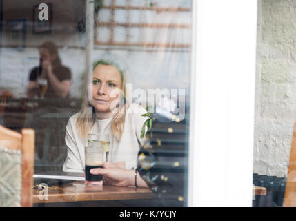 Pensive blonde attractive young adult single woman in pub. Shot through window with reflections on glass