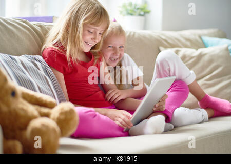 Pretty little girl looking at camera with wide smile while sitting on sofa with elder sister and watching favorite cartoon on digital tablet, interior Stock Photo