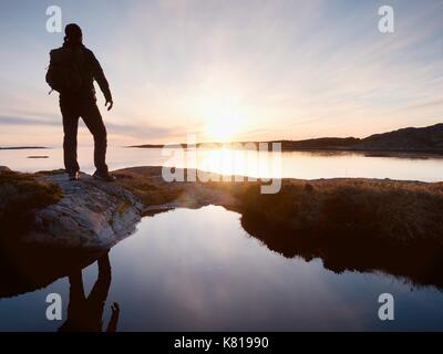 Tall backpacker watch clear sunny spring daybreak over sea. Hiker with backpack stand on rocky shore and his figure is mirrored in water pool.  Hiker  Stock Photo