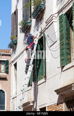 Technician standing on a ladder  on  an exterior wall installing new phone cables or lines  to a second floor apartment in an urban environment Stock Photo