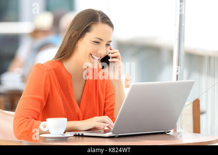 Happy customer in orange using a laptop and talking on phone sitting in a bar terrace Stock Photo