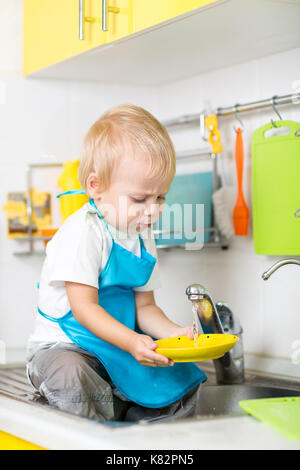 Little blond child kid boy having fun washing dishes in domestic kitchen. Stock Photo