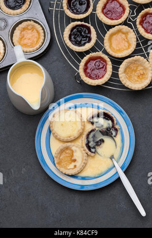 Cooked homemade jam tarts on a plate with custard next to a baking tray and a vintage circular wire rack Stock Photo