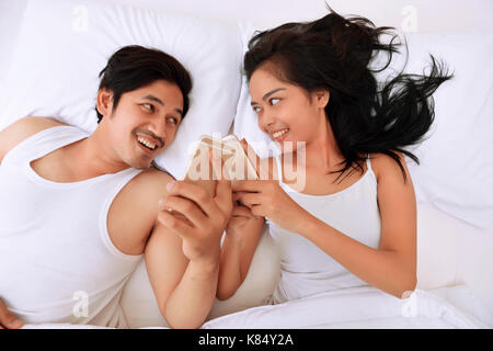 Young asian couple lying down in bed and using mobile phones at home Stock Photo