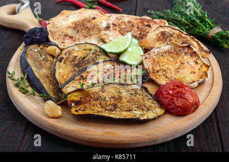 Baked vegetables: eggplant, patisson, tomatoes on a round wooden stand on a dark background. Vegan dish.