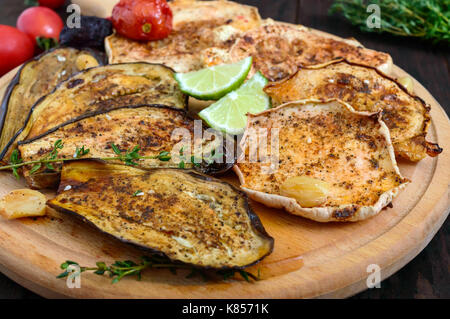 Baked vegetables: eggplant, patisson, tomatoes on a round wooden stand on a dark background. Vegan dish.