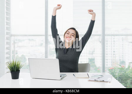 Happy relaxed young woman sitting at her desk with a laptop, stretching her arms Stock Photo