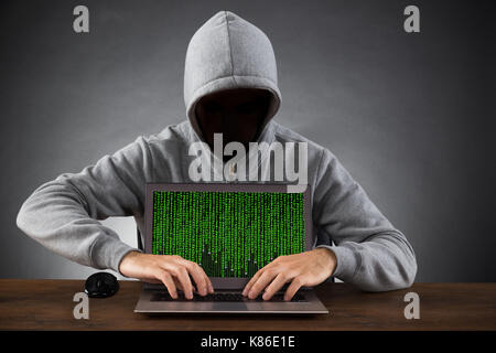 Man In Hood And Mask Hacking Laptop At Wooden Desk Stock Photo