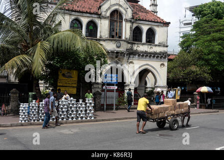 A market trader pulling is barrow loaded with goods up a slope in Pettah Market in Colombo, Sri Lanka.   On the pavement is a stack of cooking pots.   Stock Photo