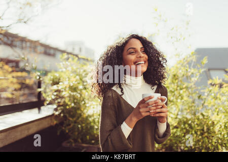 Young woman drinking coffee outdoors. Smiling curly haired woman holding a coffee cup. Stock Photo