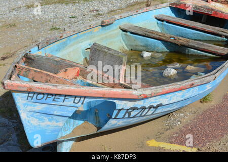 Old painted rowing boat with hole in side filled with water and rocks on dry land on promenade close to the beach at Morecambe bay England sand clouds Stock Photo