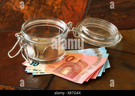 Euro coins in a jar with Euro banknotes underneath; shown on a tiled table top Stock Photo