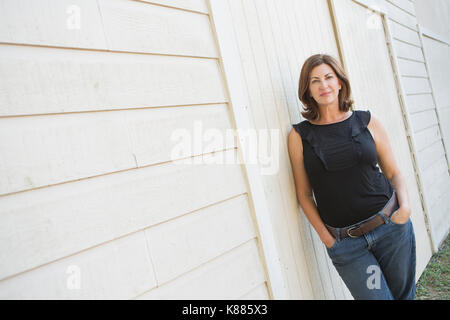 A mature woman in a sleeveless shirt with shoulder length brunette hair, hands in her pockets, leaning against a white painted wall. Stock Photo