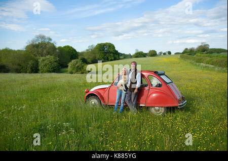 Man and woman standing side by side on a meadow, holding small dog, leaning against red vintage car. Stock Photo