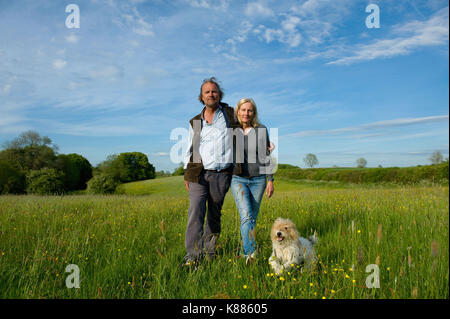 Man and woman walking arm in arm across a meadow, small dog running beside them. Stock Photo