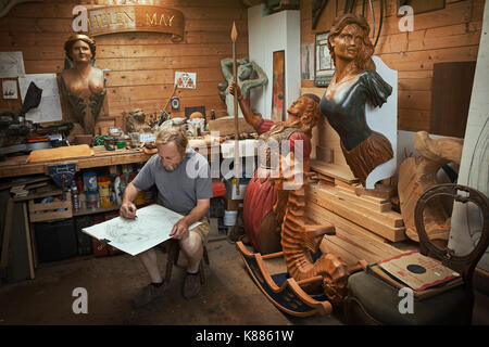A craftsman, woodworker seated on a stool in a workshop working on a drawing, sketching using charcoal. Surrounded by wooden carved and painted female Stock Photo