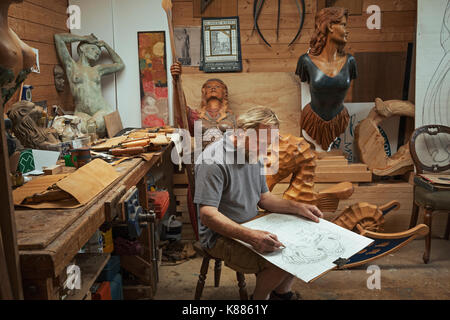 A craftsman, woodworker seated on a stool in a workshop working on a drawing, sketching using charcoal. Surrounded by wooden carved and painted female Stock Photo