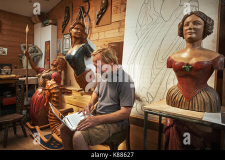 A woodworker, skilled craftsman sitting in a workshop using a laptop on his knees, surrounded by carved wooden female ship's figureheads. Stock Photo