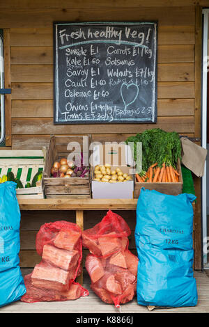 Handwritten blackboard on wall and crates with fresh vegetables and stack of firewood in red net bags outside a farm shop. Stock Photo