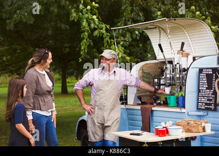 Bearded man wearing apron, woman and girl standing by blue mobile coffee shop, smiling and chatting. Stock Photo