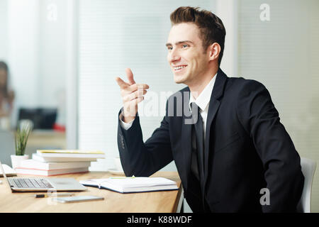 Cheerful young entrepreneur with toothy smile pointing at something while sitting in front of modern laptop at office, waist-up portrait Stock Photo