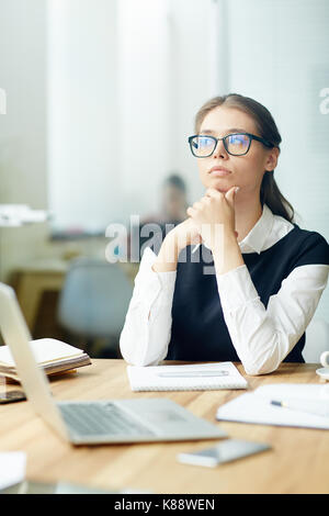 Beautiful young manager rubbing chin and looking away pensively while thinking over promising project, interior of open plan office on background Stock Photo