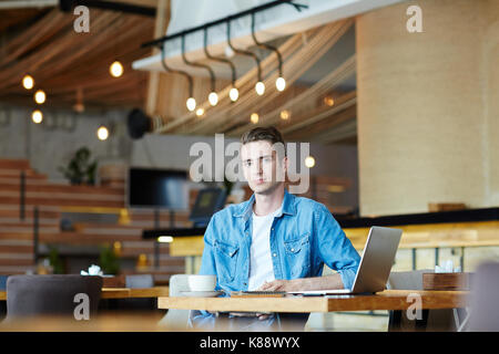 Waist-up portrait of handsome young designer looking at camera while working on laptop at modern coffeehouse, blurred background Stock Photo