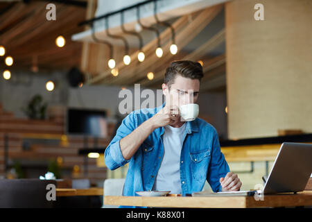 Young manager with stylish haircut enjoying fragrant coffee while working on promising project with help of laptop, interior of spacious cafe on backg Stock Photo