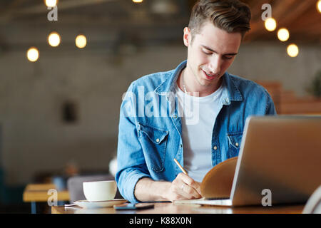 Confident young manager wearing denim shirt sitting at cafe table and writing down creative ideas in notepad, blurred background Stock Photo