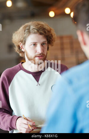 Waist-up portrait of bearded red-haired manager having project discussion with his colleague and taking necessary notes, blurred background Stock Photo