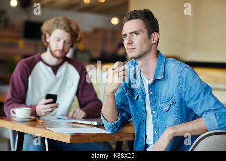 Handsome young manager looking away pensively while sitting at cafe table with his bearded colleague and working on start-up project, blurred backgrou Stock Photo