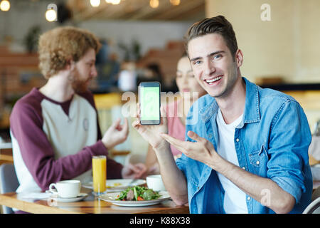 Handsome young man with wide smile posing for photography while pointing at blank screen of modern smartphone, his friends chatting animatedly with ea Stock Photo