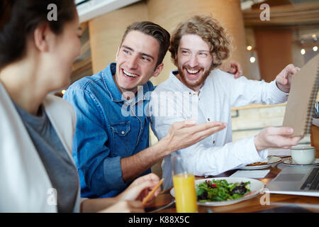 Joyful group of managers having fun while working together on joint project at cozy small cafe, two handsome men showing notepad with their ideas to s Stock Photo