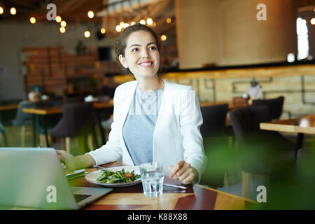Attractive young financial manager with charming smile looking away while preparing annual accounts on laptop and enjoying delicious lunch at modern r Stock Photo
