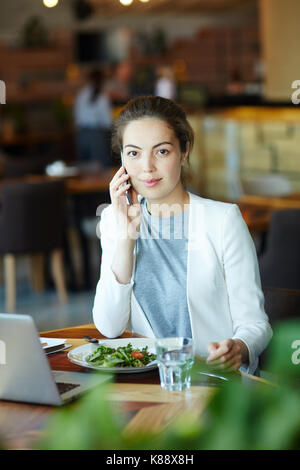 Waist-up portrait of confident young entrepreneur looking at camera with small smile while conducting telephone negotiations at cozy small cafe, blurr Stock Photo