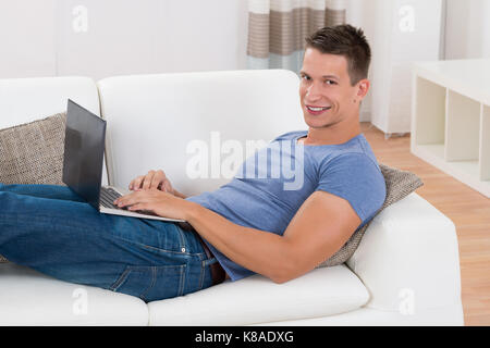 Young Man Smiling While Using Laptop In Living Room Stock Photo