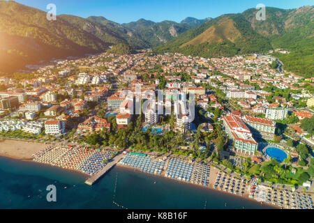 Aerial view of beautiful city, sandy beach with chaise-lounges, mountains, green trees, hotels, buildings, pool, at sunset in Icmeler, Turkey. Summer  Stock Photo