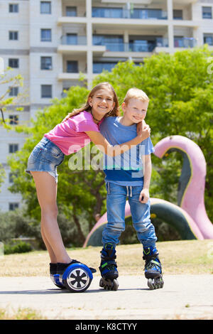 Girl on hoverboard with her brother on rollers having fun outdoors Stock Photo