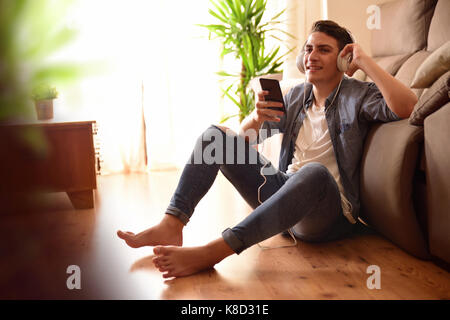 Teen listening and music with headphones from mobile sitting on the floor leaning on a couch in the living room Stock Photo