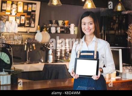 Smiling asian woman barista holding blank tablet compute in front of coffee shop counter bar,Mock up space for display of menu or design,clipping path Stock Photo