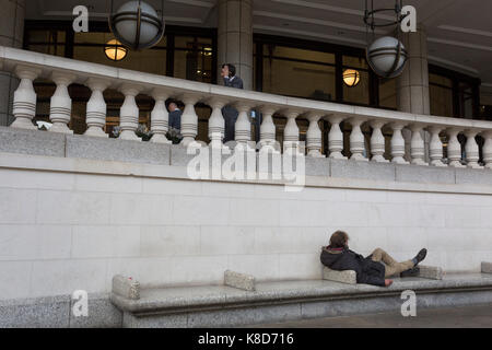 A homeless man lies on City seating as a businessman smokes above, on 14th September 2017, in the City of London, England. Stock Photo