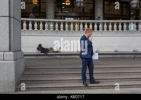 A homeless man lies on City seating as a businessmen smokes, on 14th September 2017, in the City of London, England. Stock Photo